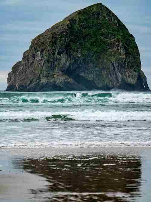 A surfer rides the waves a way off in the distance with a large rock behind them